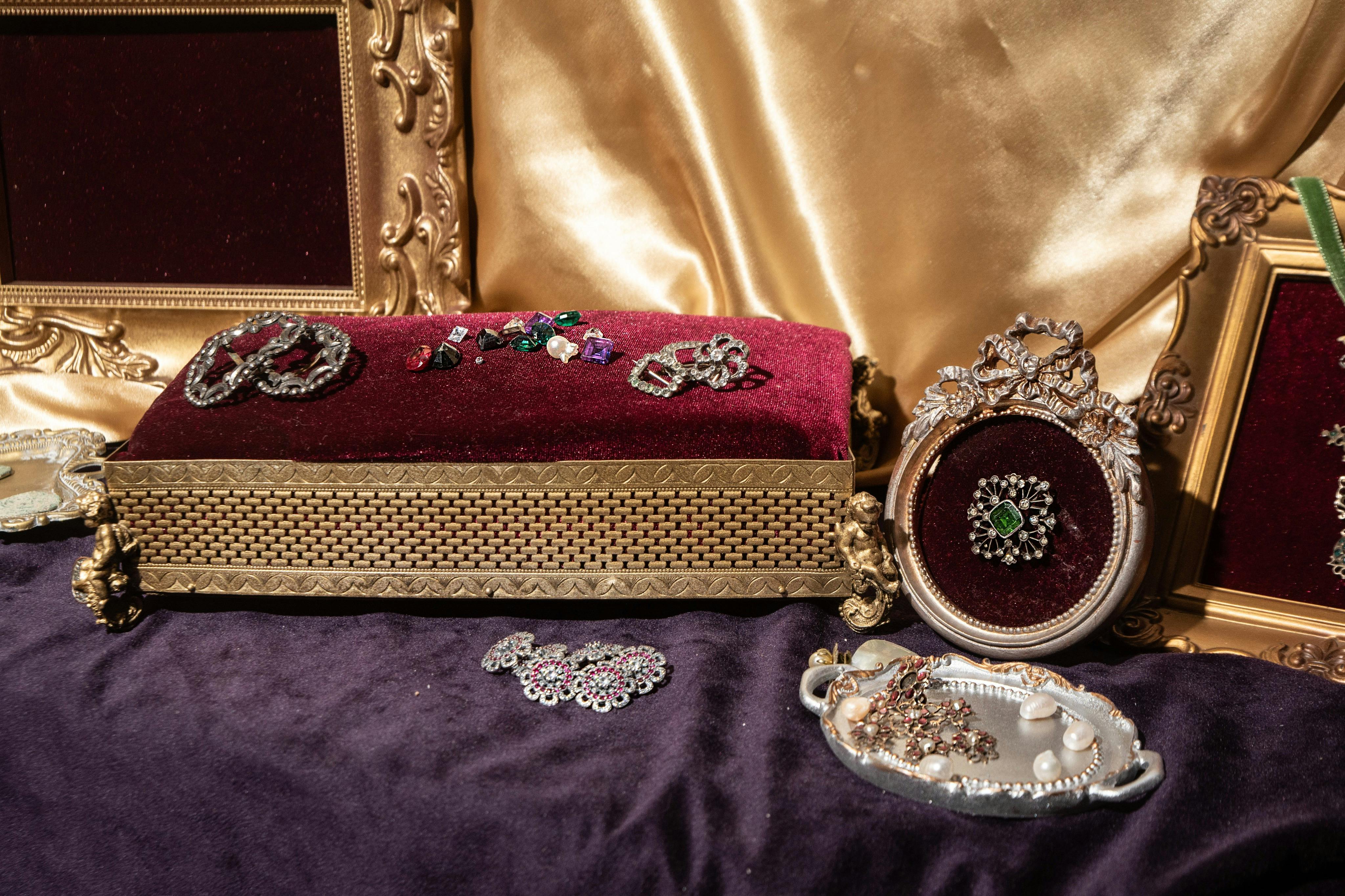 Ozdobná krabička s červeným sametovým víkem pokrytým šperky, obklopená zarámovanými uměleckými díly a podnosem s perlami a náhrdelníkem.