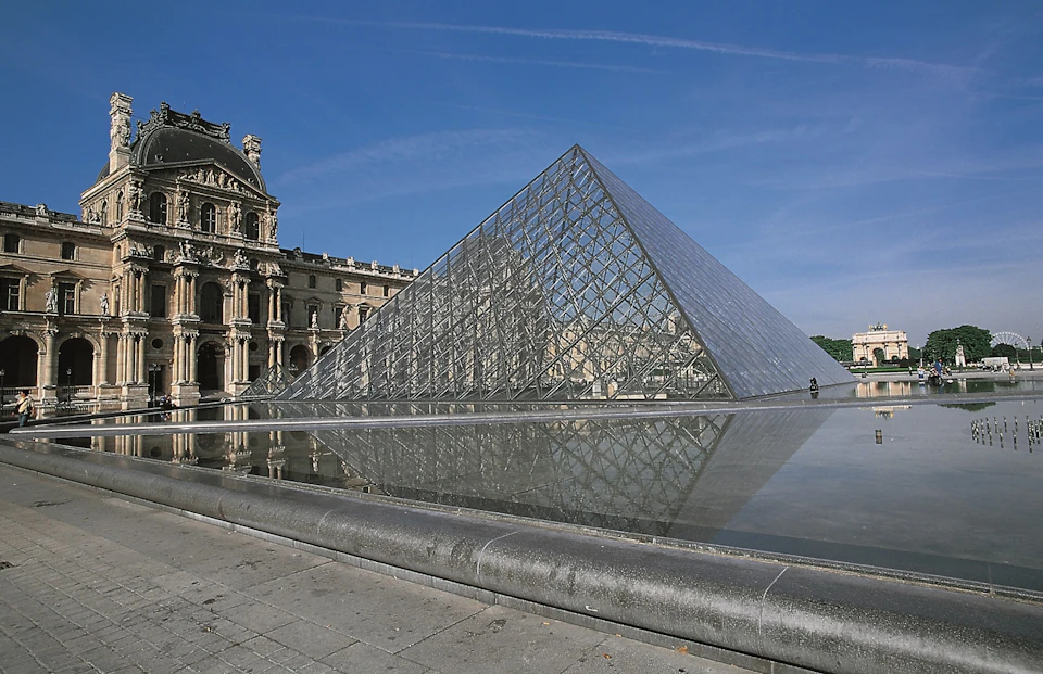 Glass pyramid with a water reflection next to historic building under a clear blue sky.