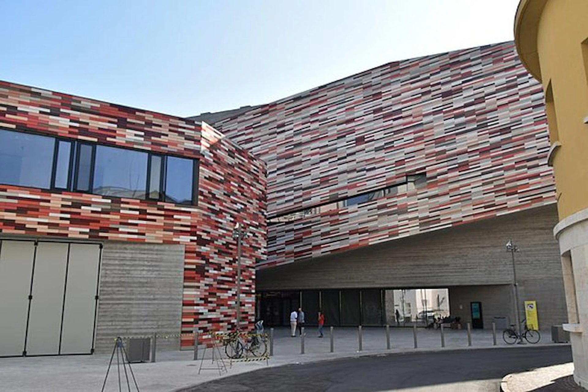 Modern building with a colorful, patterned façade, large windows, and an open entrance. Three people and several bicycles are visible.
