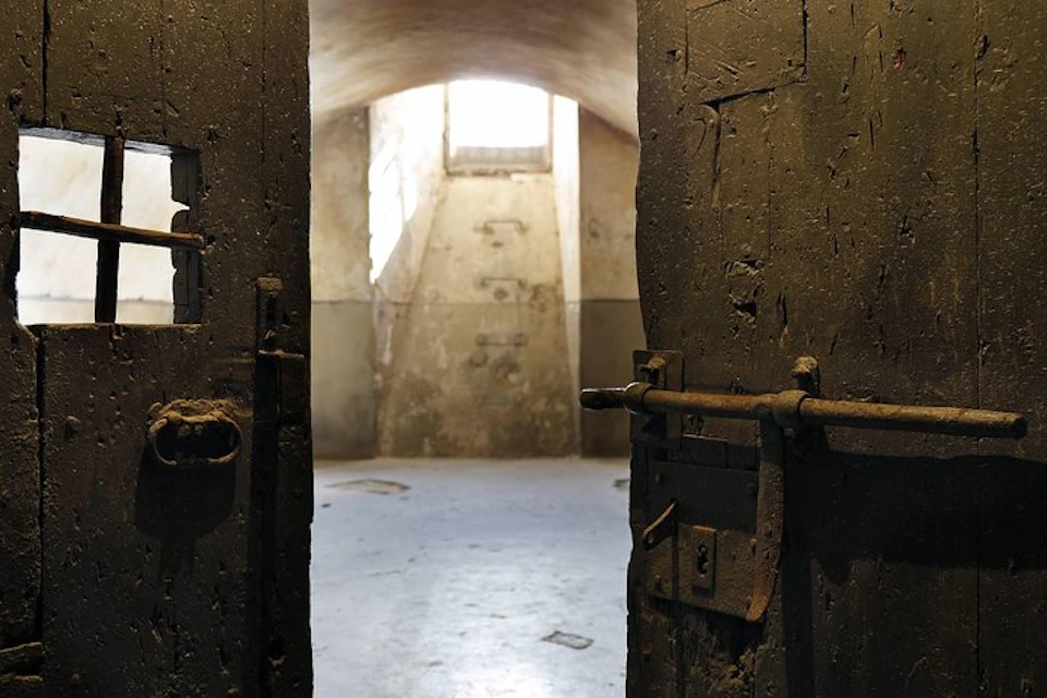 A dimly lit, empty room with a vaulted ceiling and a small, barred window, viewed through open, rusty metal doors.