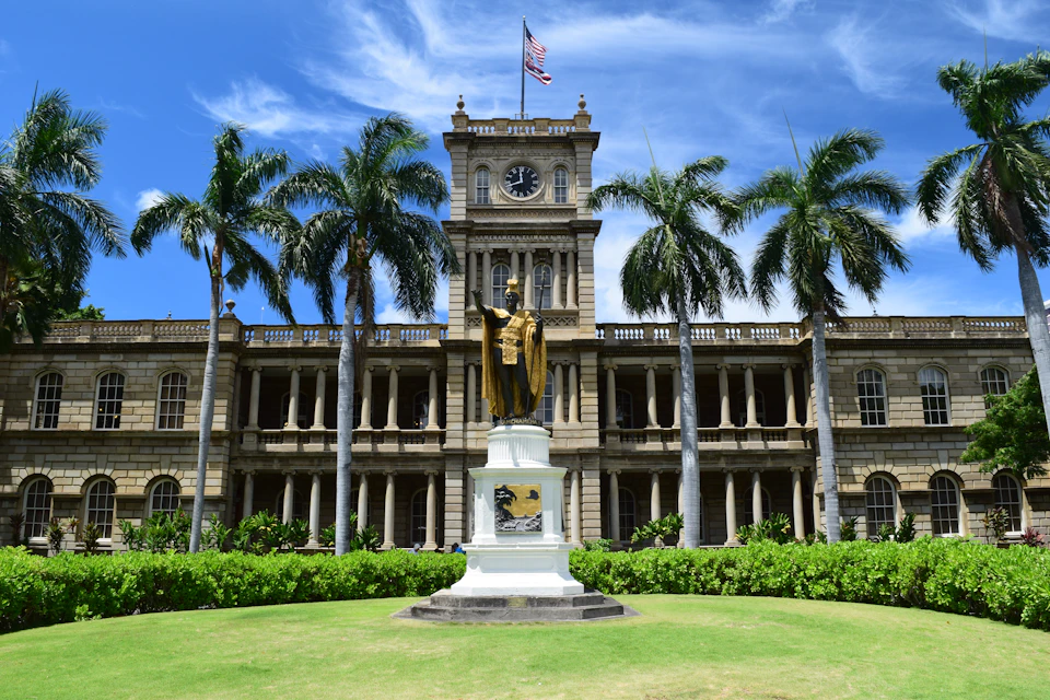 Ornate historical building with a central clock tower and American flag. Statue of a person in gold robes stands in front. Palm trees line the scene.