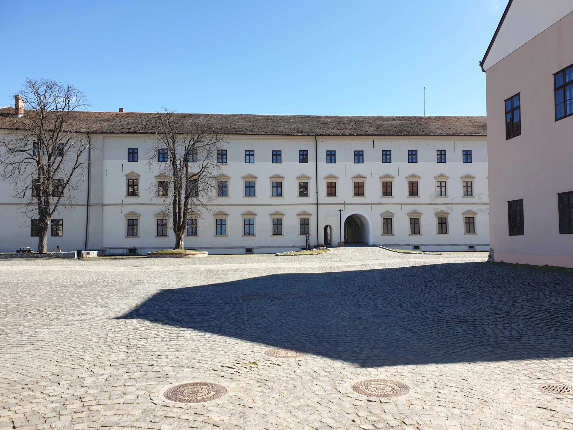 A large, white building with multiple windows and an archway entrance, set in a cobblestone courtyard under a clear blue sky.