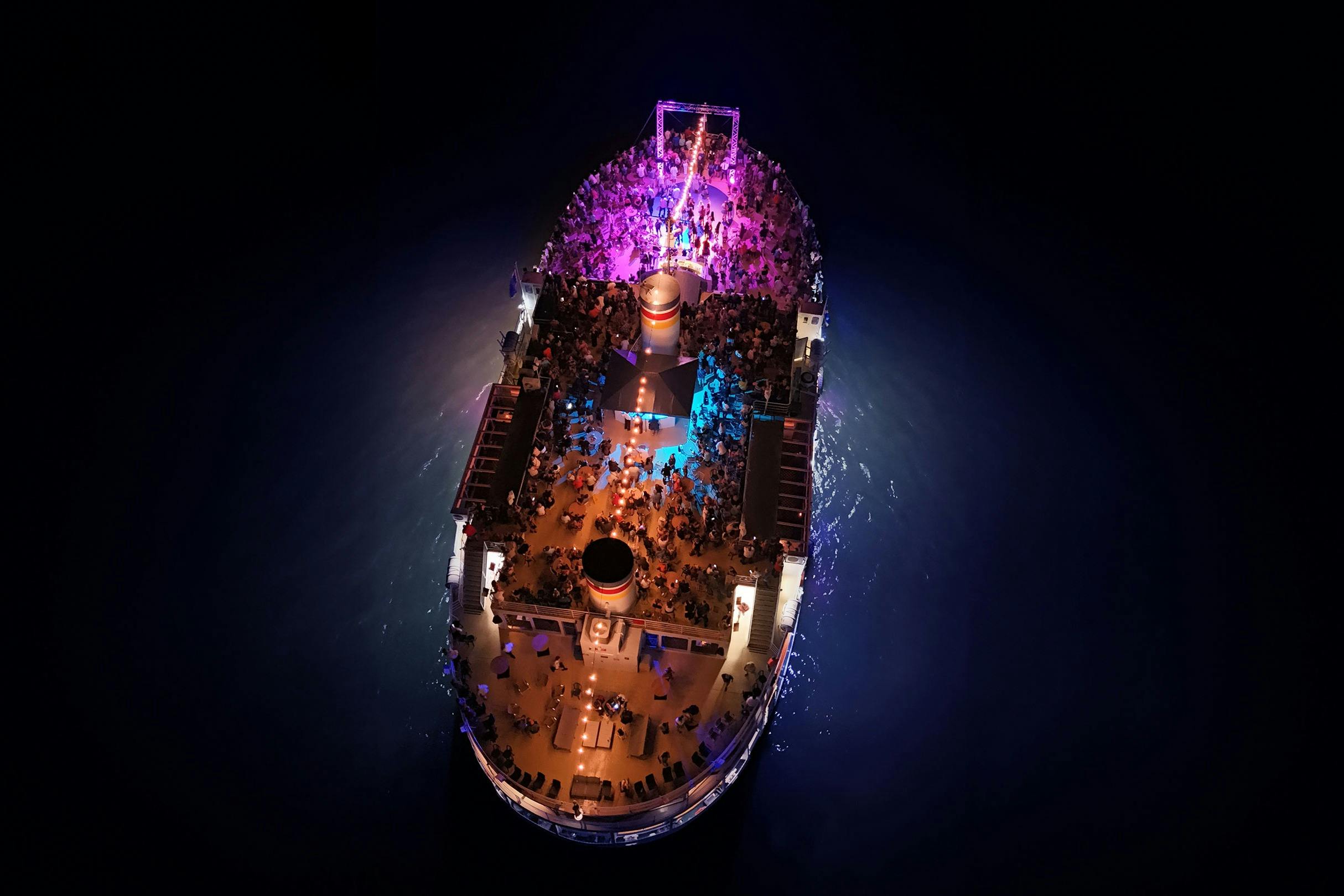 Vue aérienne d'un bateau de croisière de nuit, illuminé de lumières colorées, avec de nombreuses personnes rassemblées sur son pont.
