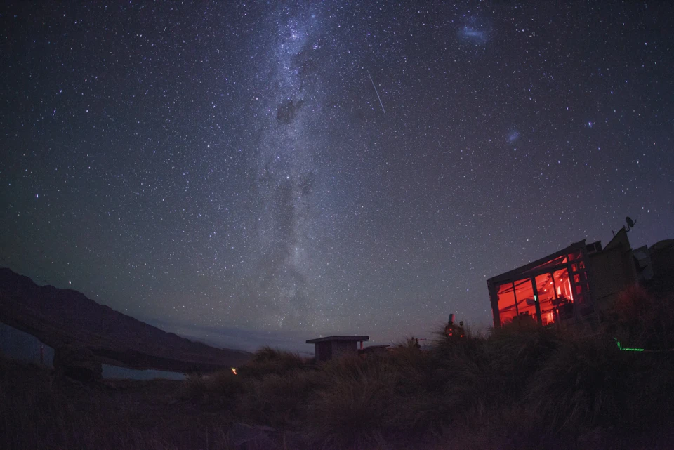 Starry night sky with the Milky Way visible, a streaking meteor, and a lit building with red light on the right.