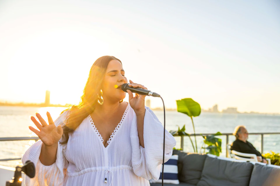 A woman sings into a microphone outdoors at sunset, with water and distant buildings in the background. An older person sits nearby.
