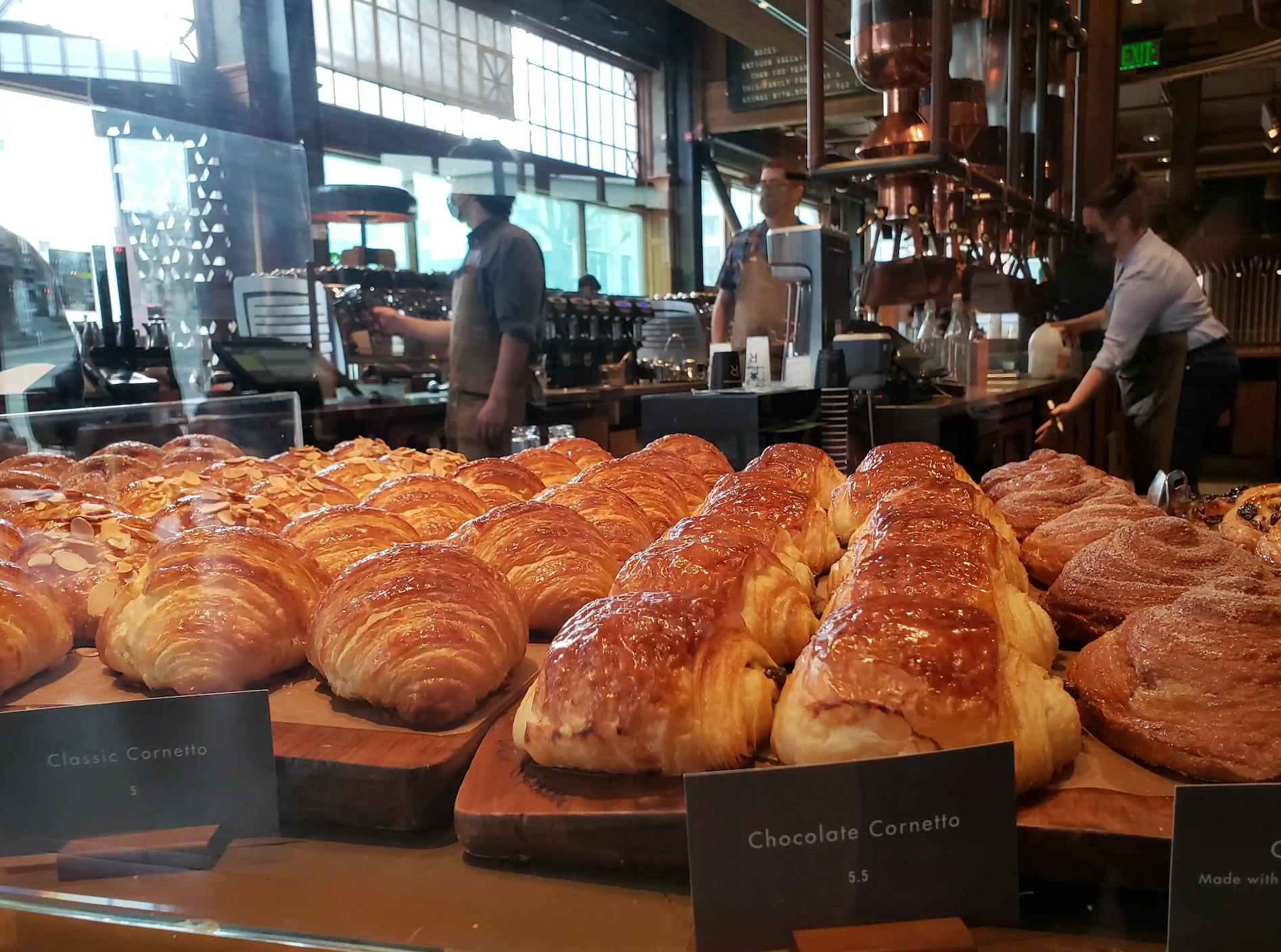 Filas de cruasanes y cornettos de chocolate en una vitrina de panadería; los baristas trabajan al fondo.