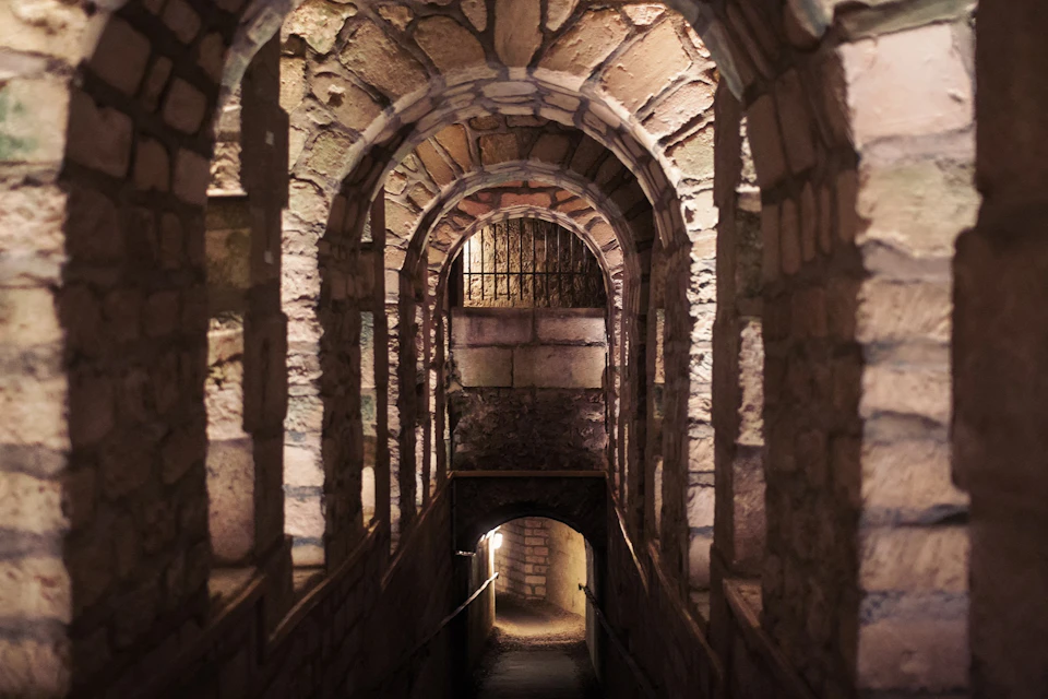 A stone hallway with arched ceilings and warm lighting, leading to a gated opening at the end.