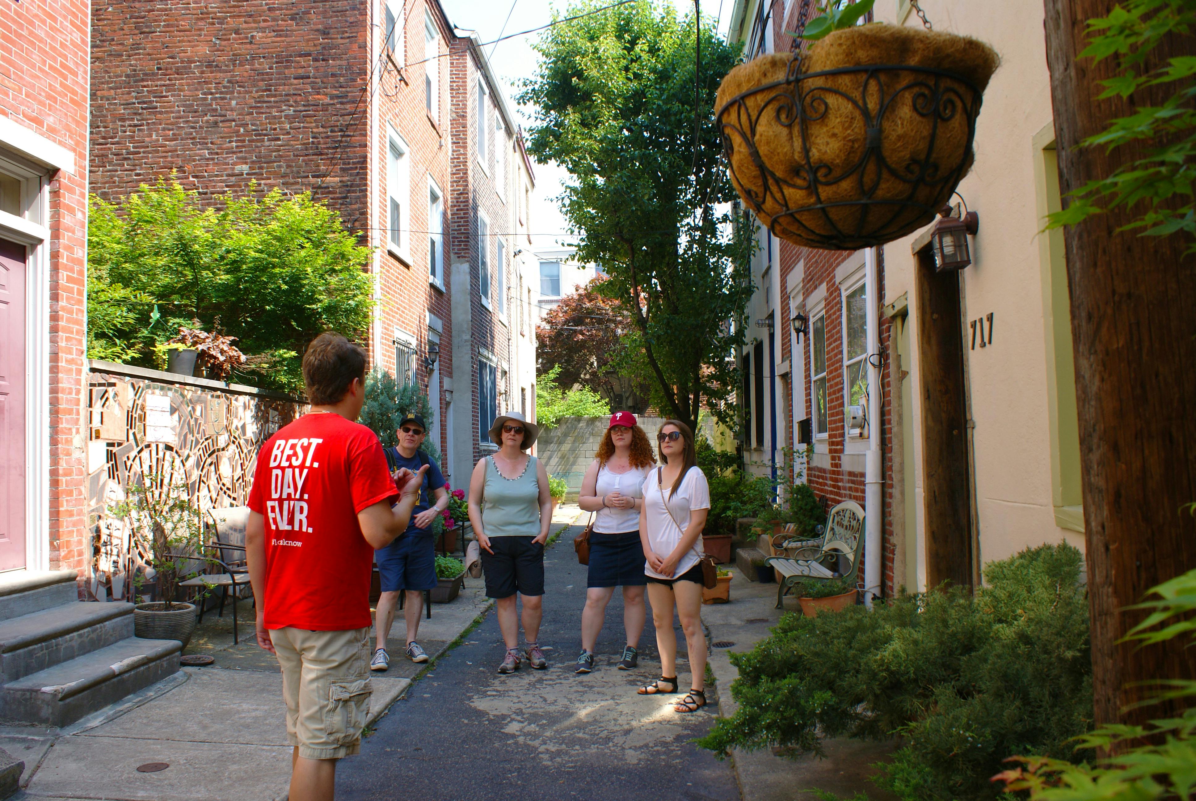 Een rondleider praat met vier mensen in een steeg met bakstenen gebouwen en groen.