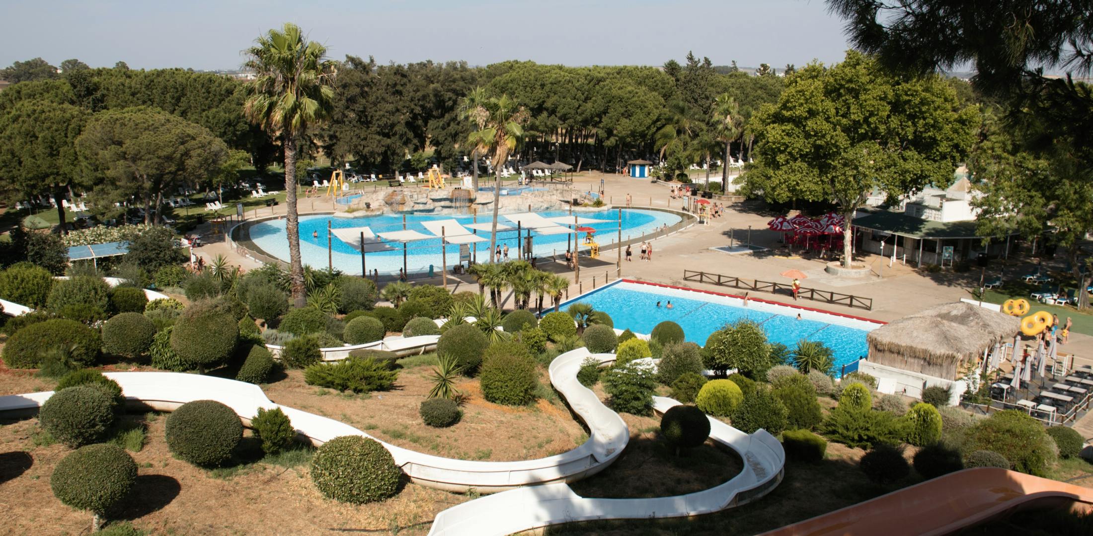 Un parc aquatique avec des piscines, des toboggans, des palmiers et des places assises ombragées sous un ciel clair entouré de verdure.