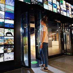 En person i avslappnad klädsel står nära en svängdörr, omgiven av en vägg med många färgglada bildskärmar.