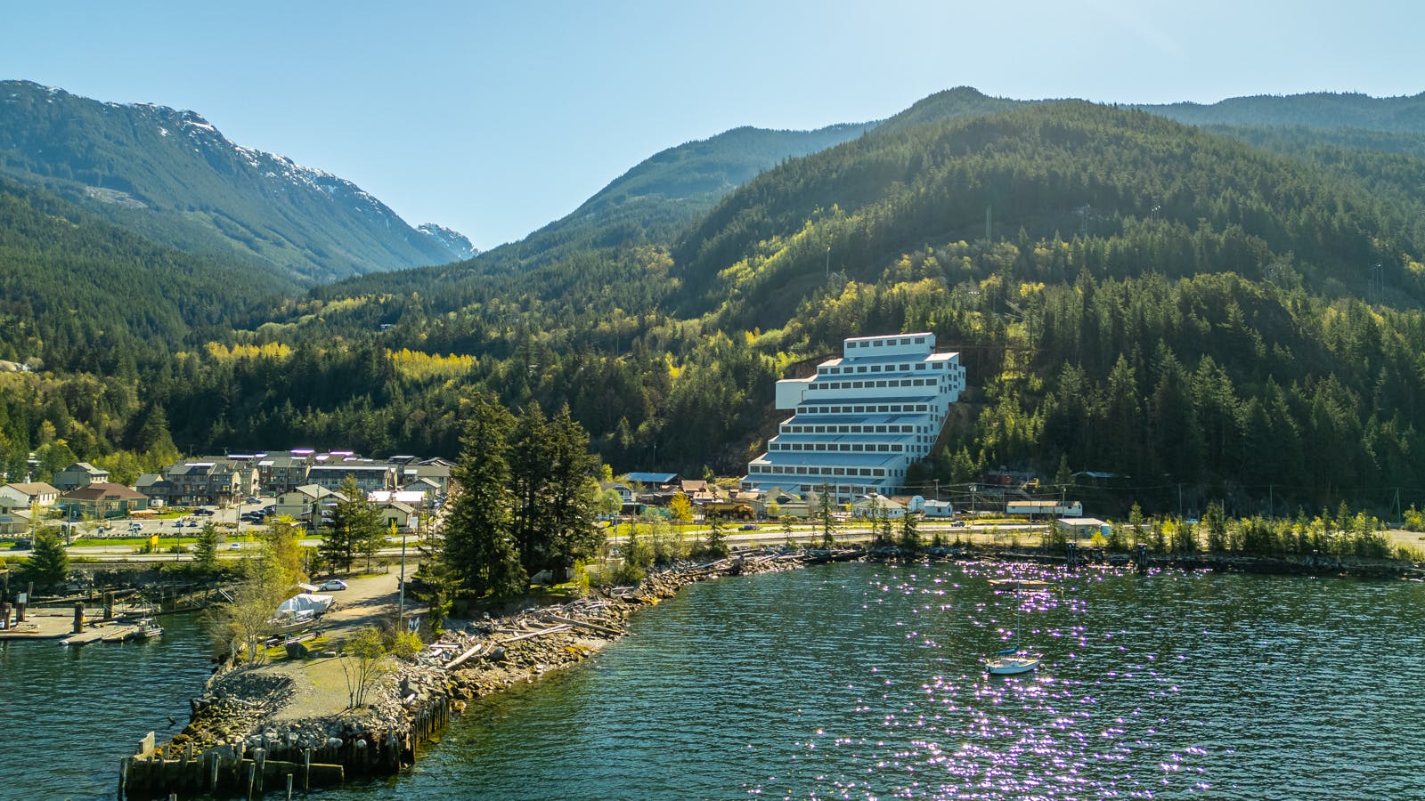 A tiered white building is set against a backdrop of forested mountains, with a lake and a sailboat in the foreground.