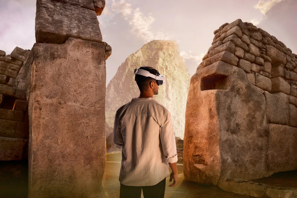 A person wearing virtual reality goggles stands between two large stone structures, with a mountain peak visible in the background.