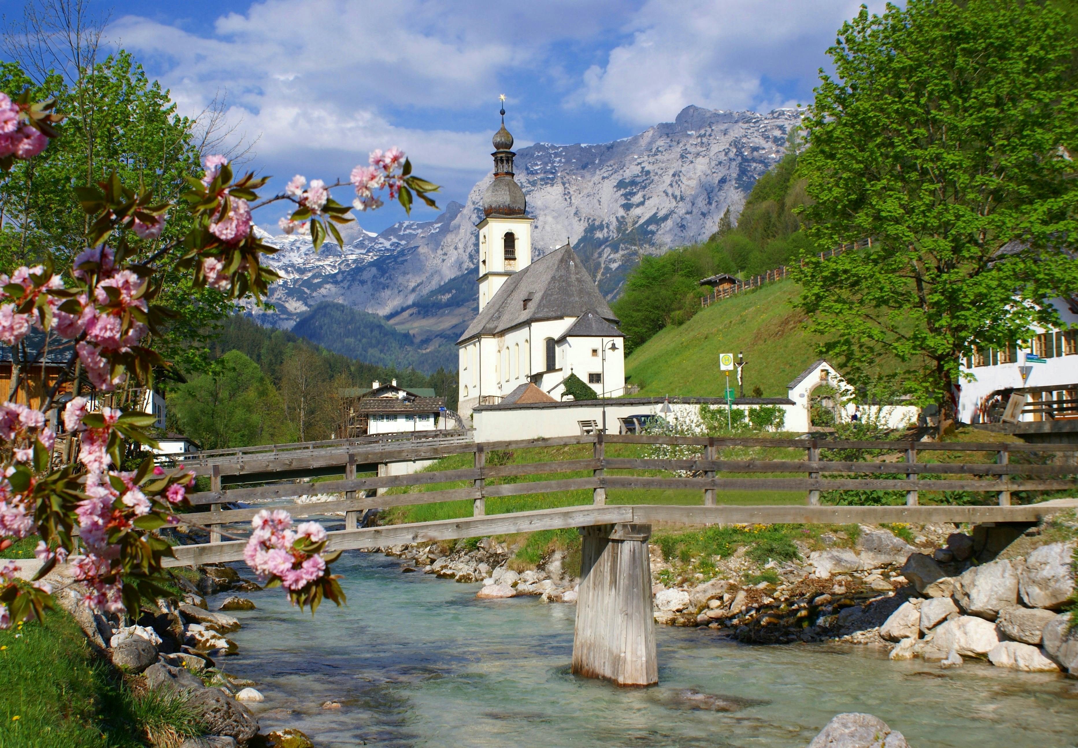 Bridge over a stream with a church and mountains in the background, surrounded by greenery and blooming flowers.