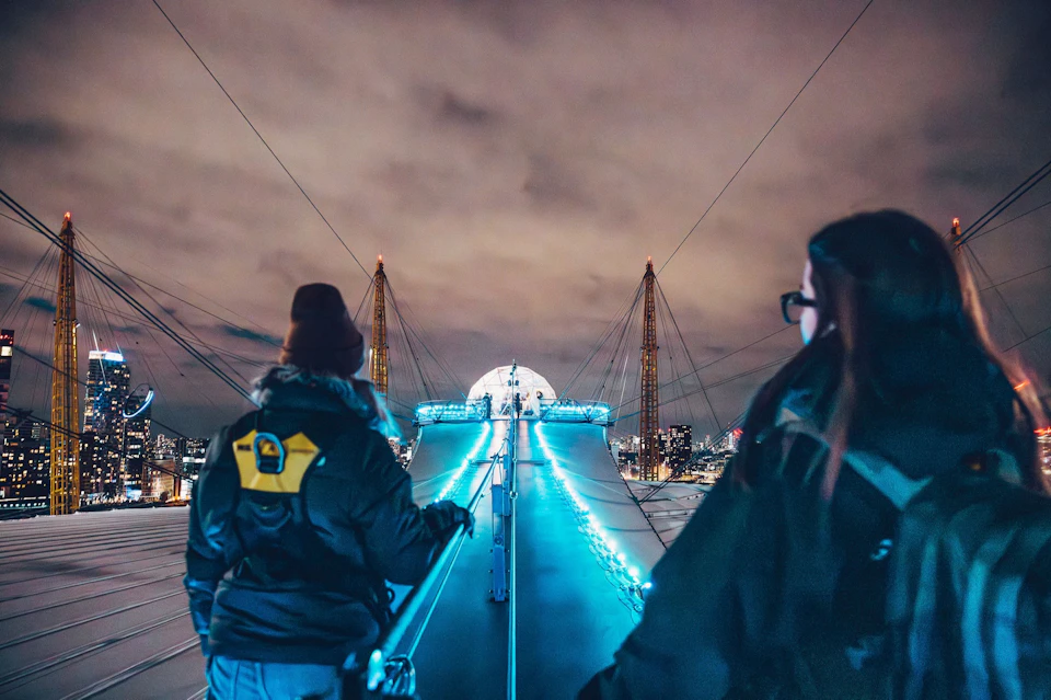Two people walking on a lit-up structure at night with city lights and a cloudy sky in the background.