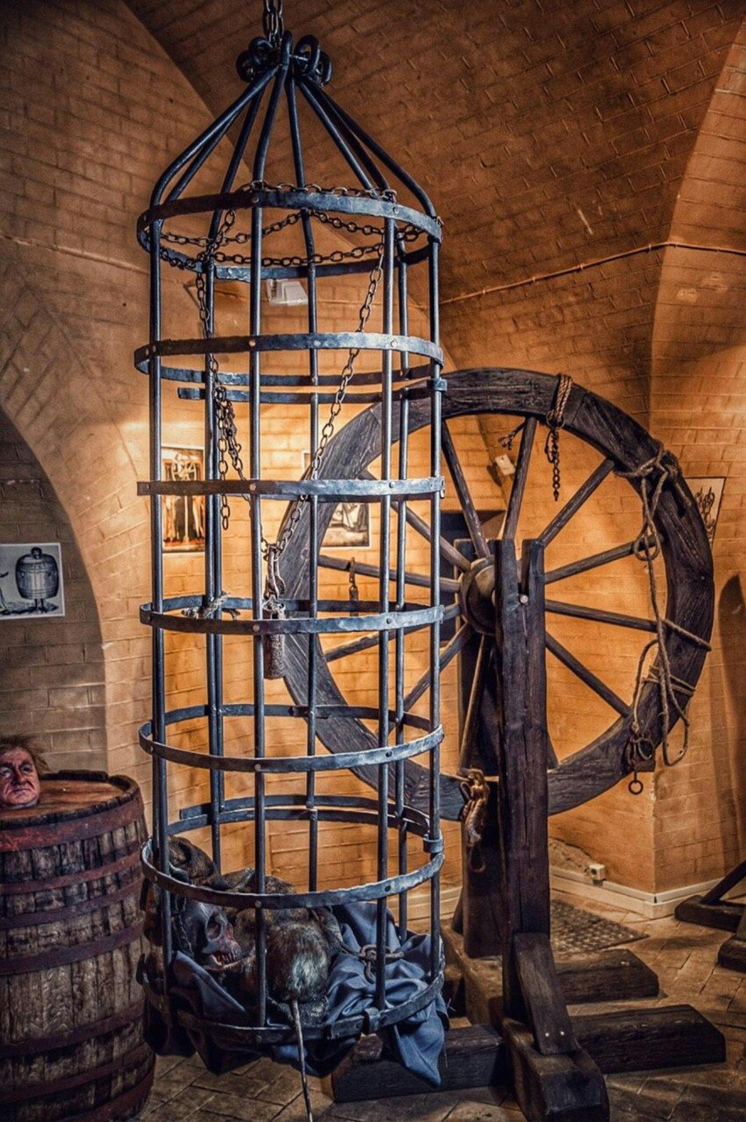 Středověká mučírna s železnou klecí, velkým dřevěným kolem a sudem s hlavou figuríny na cihlovém pozadí.