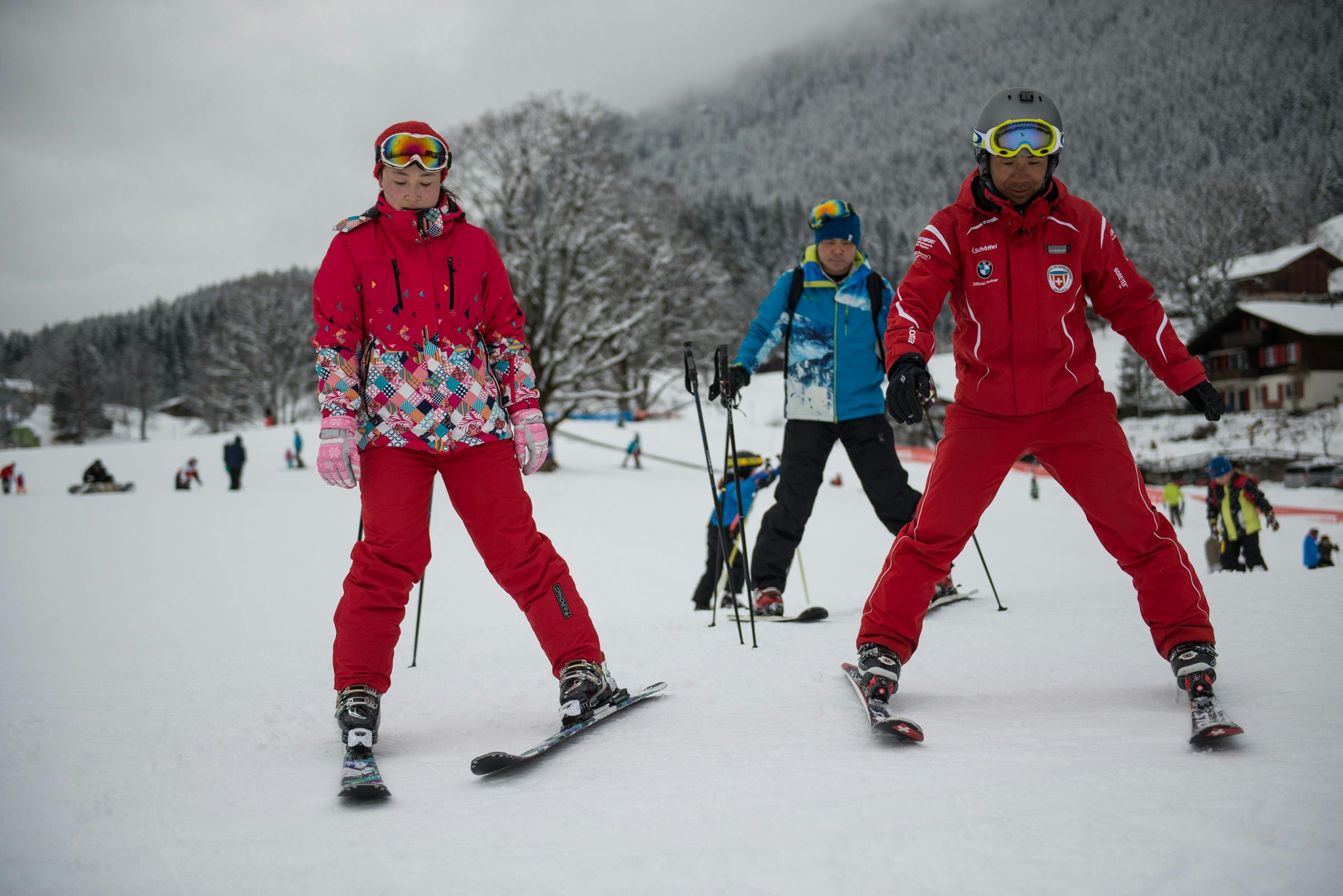 Ένα άτομο με κόκκινη στολή σκι στέκεται στα σκι σε μια χιονισμένη περιοχή, με άλλους σκιέρ και χιονισμένα δέντρα στο παρασκήνιο.