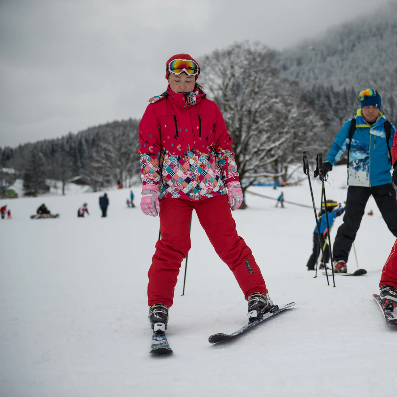 Person in a red ski outfit stands on skis in a snowy area, with other skiers and snow-covered trees in the background.