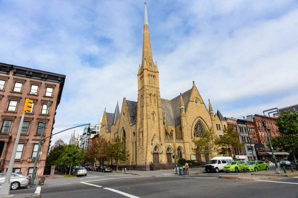 Large stone church with a tall spire on a city street corner, surrounded by parked cars and pedestrians.