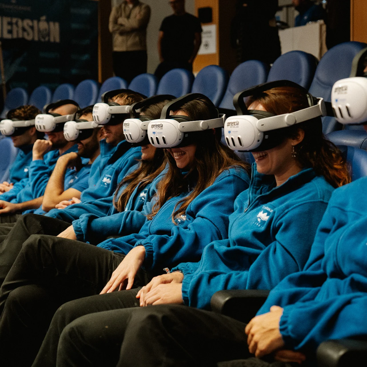 A group of people seated in rows wearing identical blue jackets and virtual reality headsets in a theater-like setting.