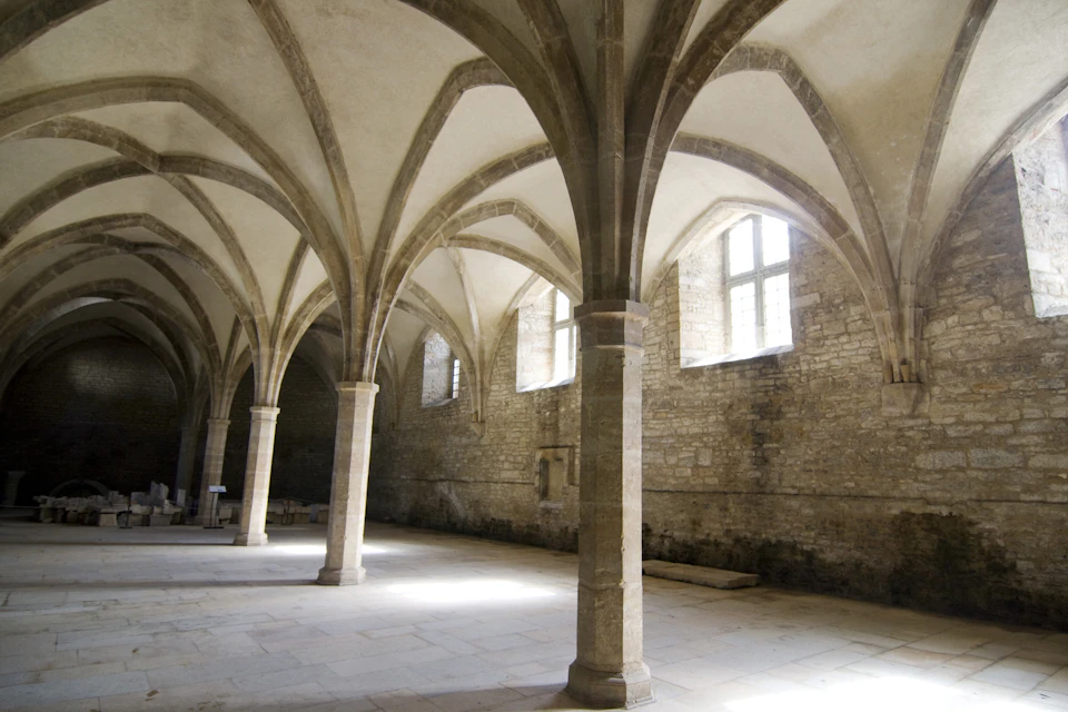 Stone hall with tall arched ceilings, several columns, and large windows. Light streams in, illuminating the space.