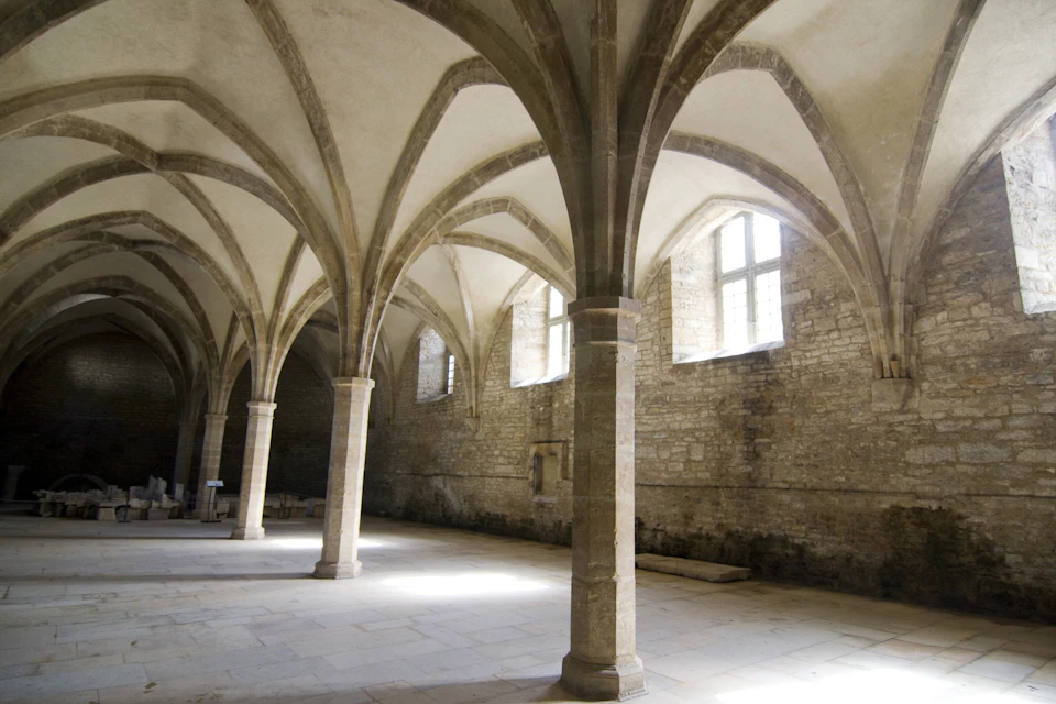 Stone hall with tall arched ceilings, several columns, and large windows. Light streams in, illuminating the space.