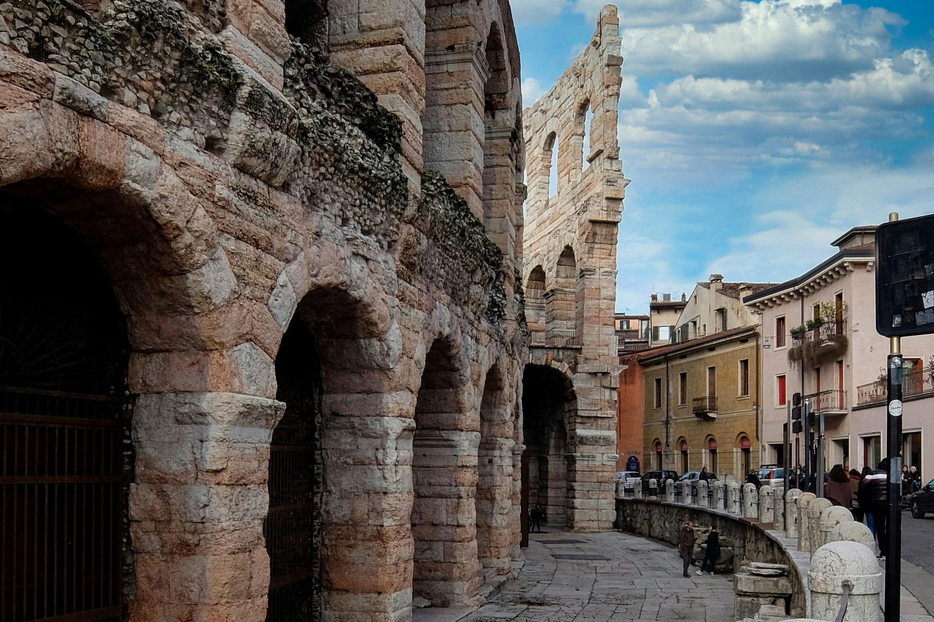 A stone amphitheater wall with arches, part of a historic structure, next to a street with buildings under a partly cloudy sky.