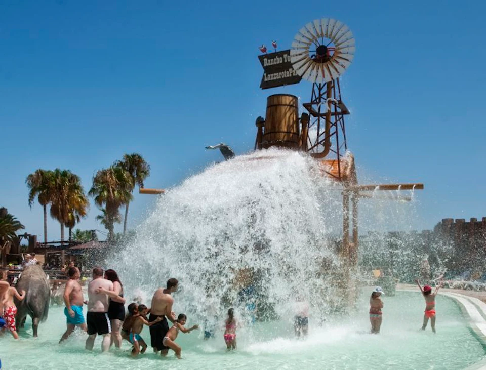 People enjoying water splashing from a large bucket under a themed structure with a windmill and palm trees in the background.
