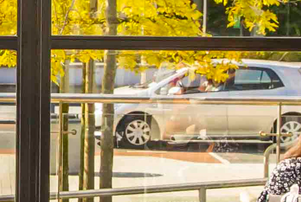 People dining on an outdoor terrace in sunny weather, surrounded by glass walls with a view of trees and buildings.