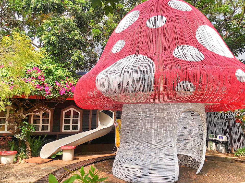 A playground with a large wire-frame mushroom structure, a slide, and a cottage-like building surrounded by greenery.