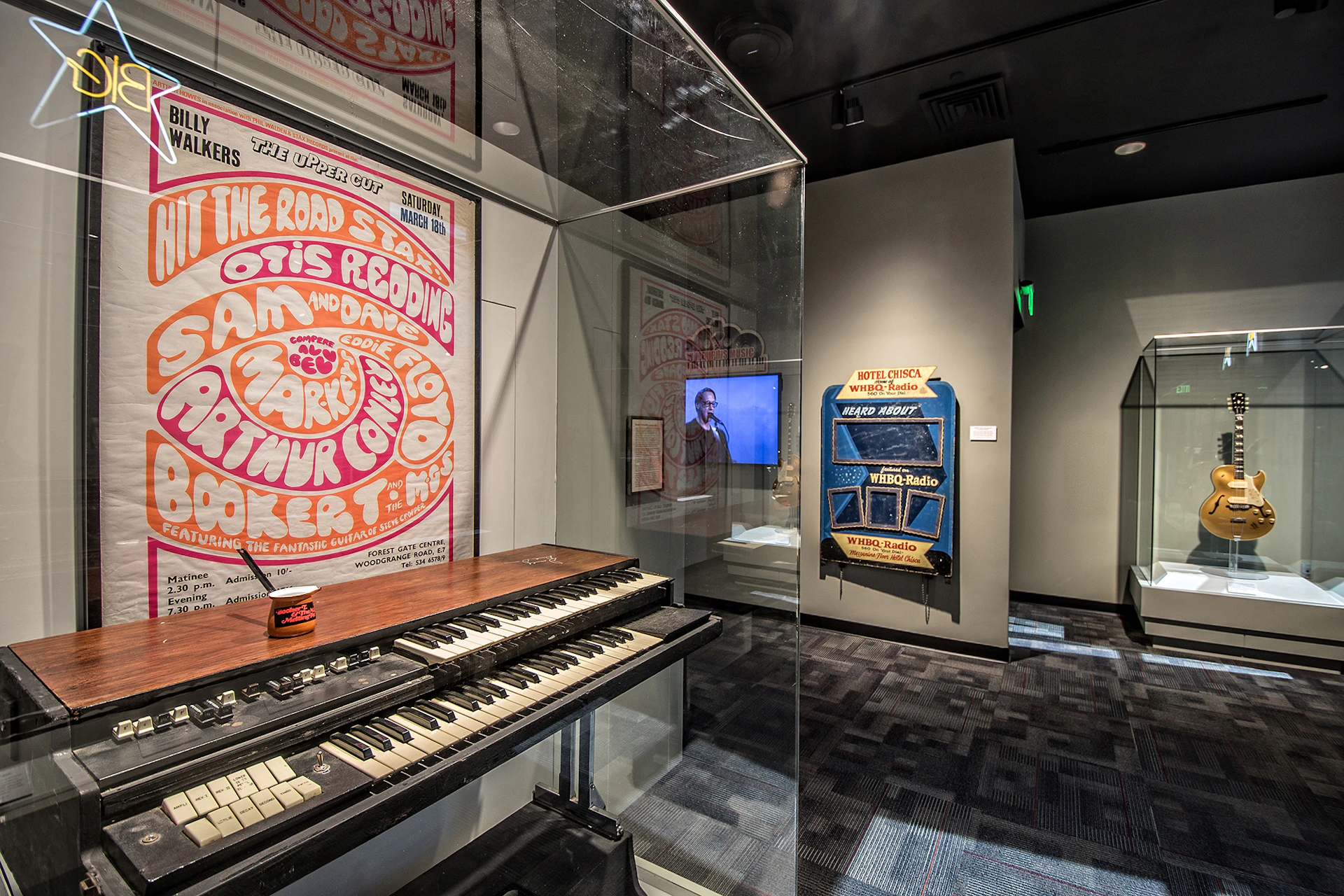 A museum exhibit featuring a vintage keyboard, a large poster, a video screen, and an old jukebox-style radio machine.
