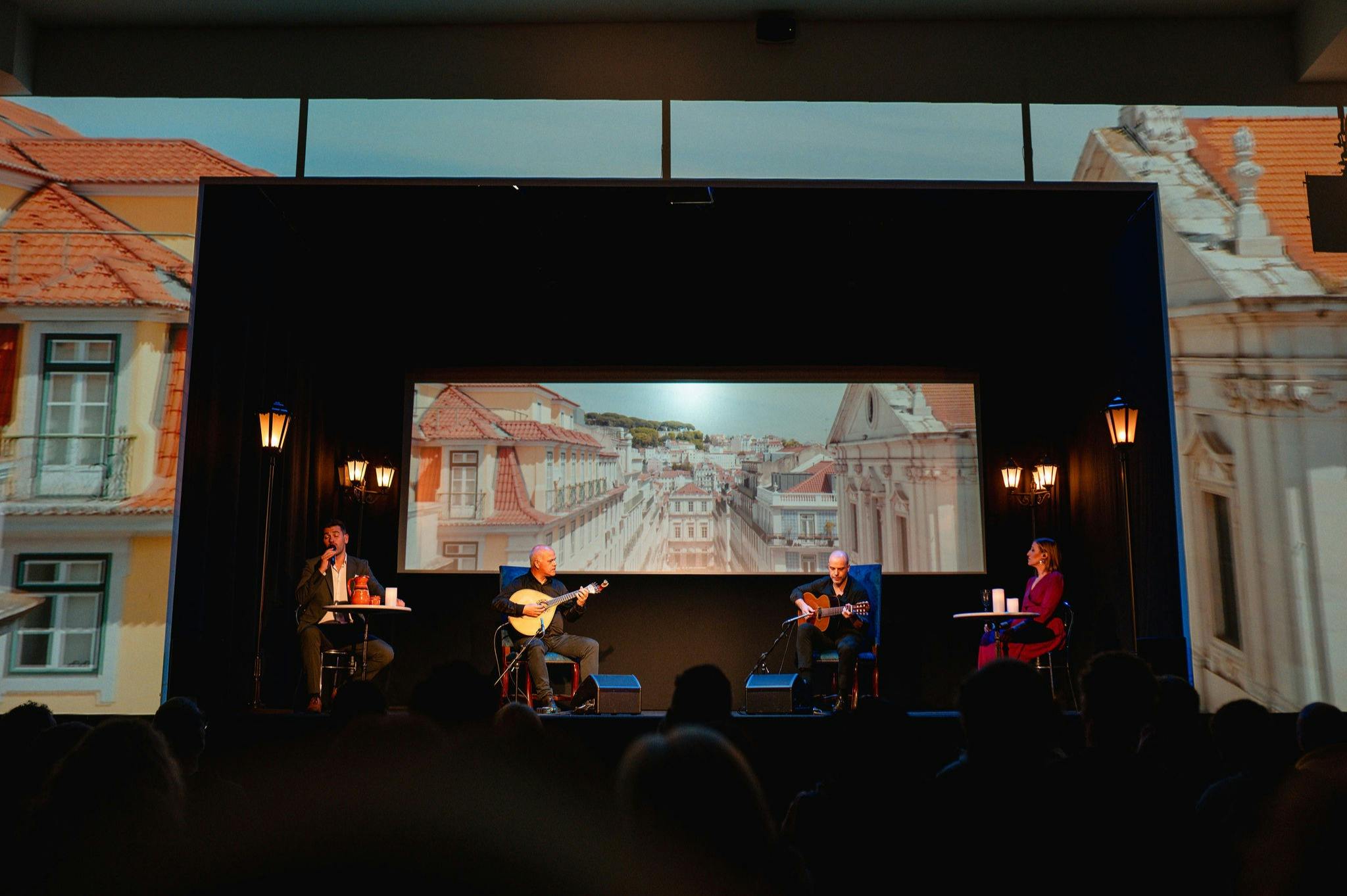 Four people are on a stage with a cityscape backdrop. Two play guitars, one reads from a book, and the other looks on.
