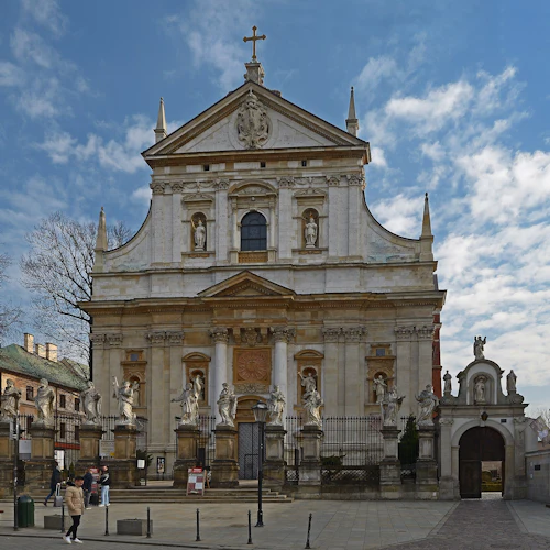 Kyrkfasad i barockstil med statyer, invecklade pelare och ett kors ovanpå. Människor som går och fotograferar framför.