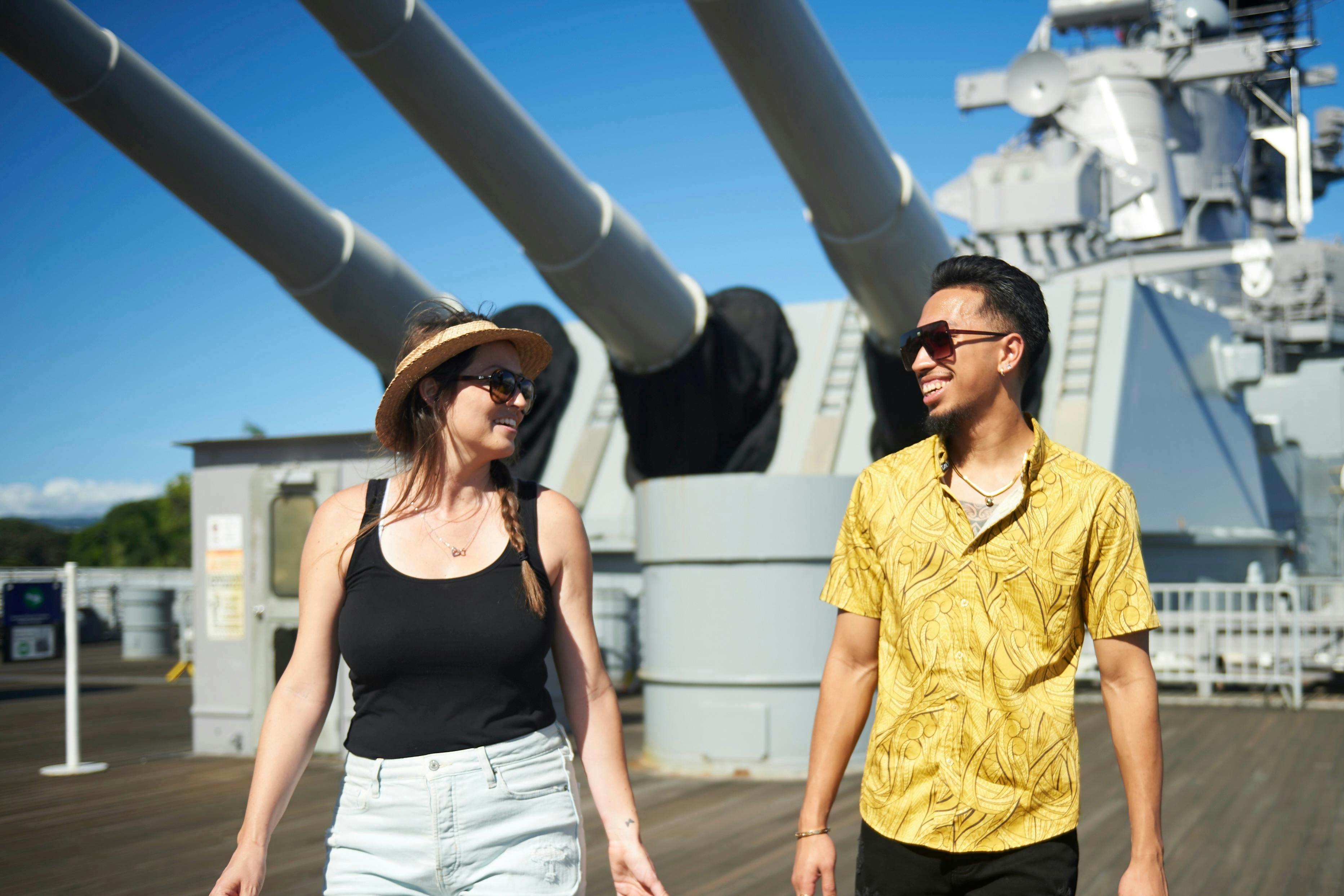Two people walking on a ship's deck with large cannons in the background, smiling under a clear sky.
