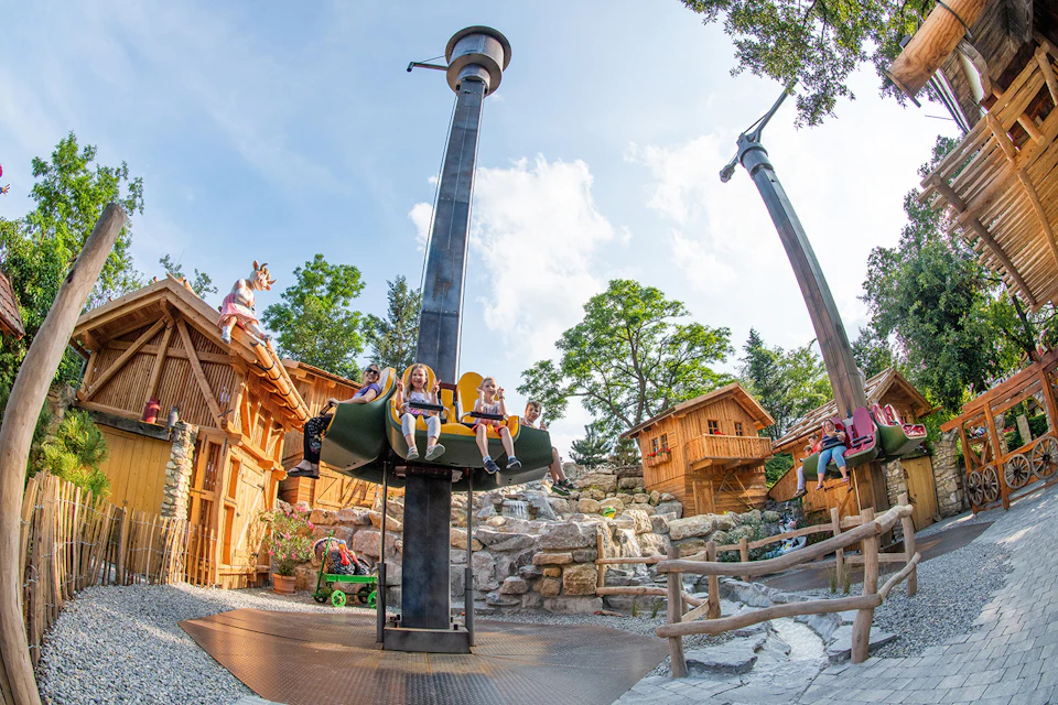 Children riding and enjoying an amusement park ride surrounded by rustic wooden buildings and rocks, under a partly cloudy sky.