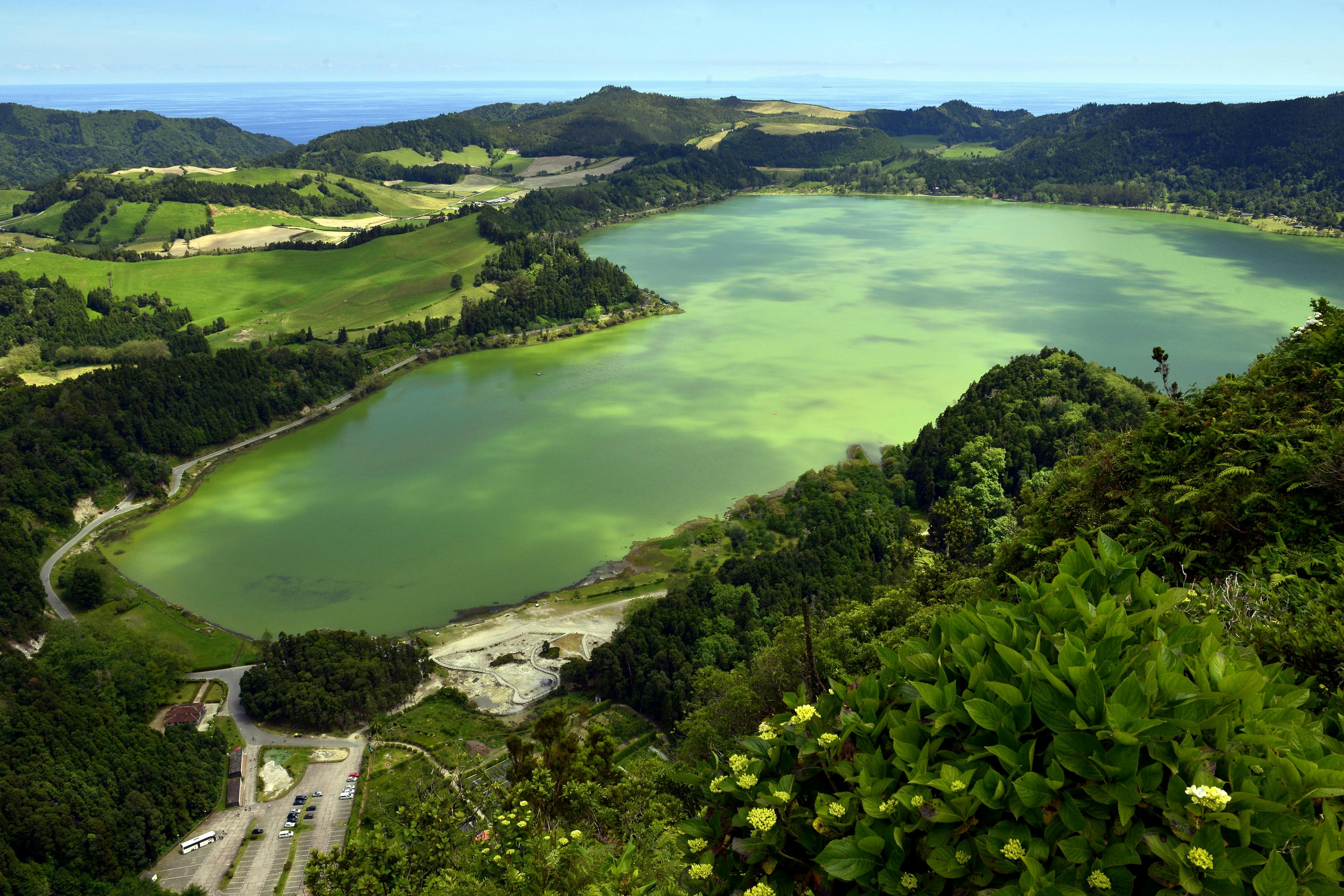 Aerial view of a green lake surrounded by lush hills, fields, trees, and a parking area with cars and a bus in the foreground.