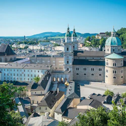 Flygfoto över en stad med historiska byggnader, kyrktorn och en katedral. Gröna kullar och blå himmel i bakgrunden.