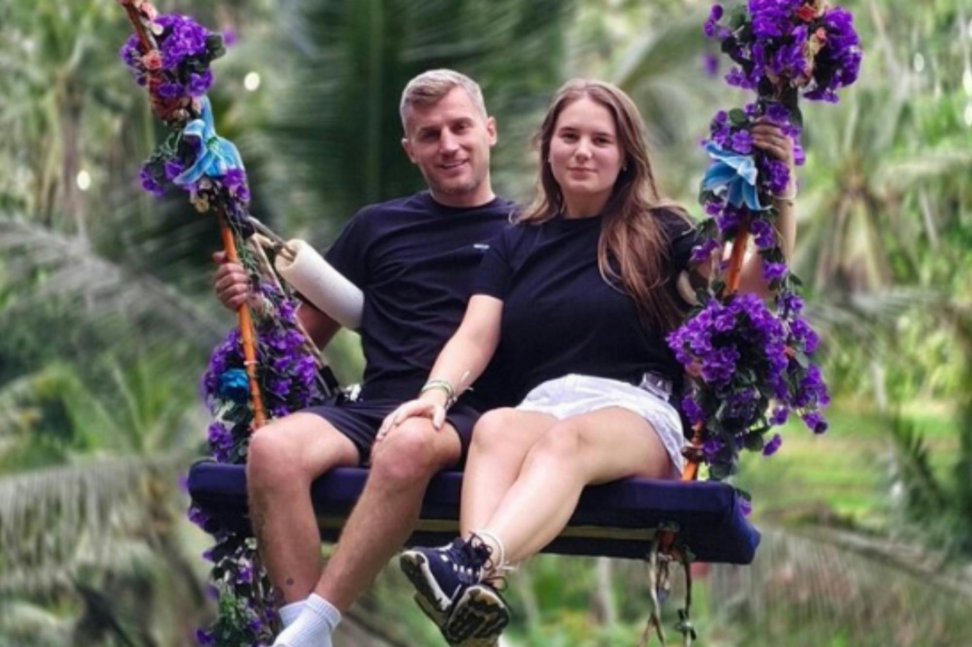 A man and a woman sitting on a swing decorated with purple flowers, in an outdoor setting with green foliage in the background.