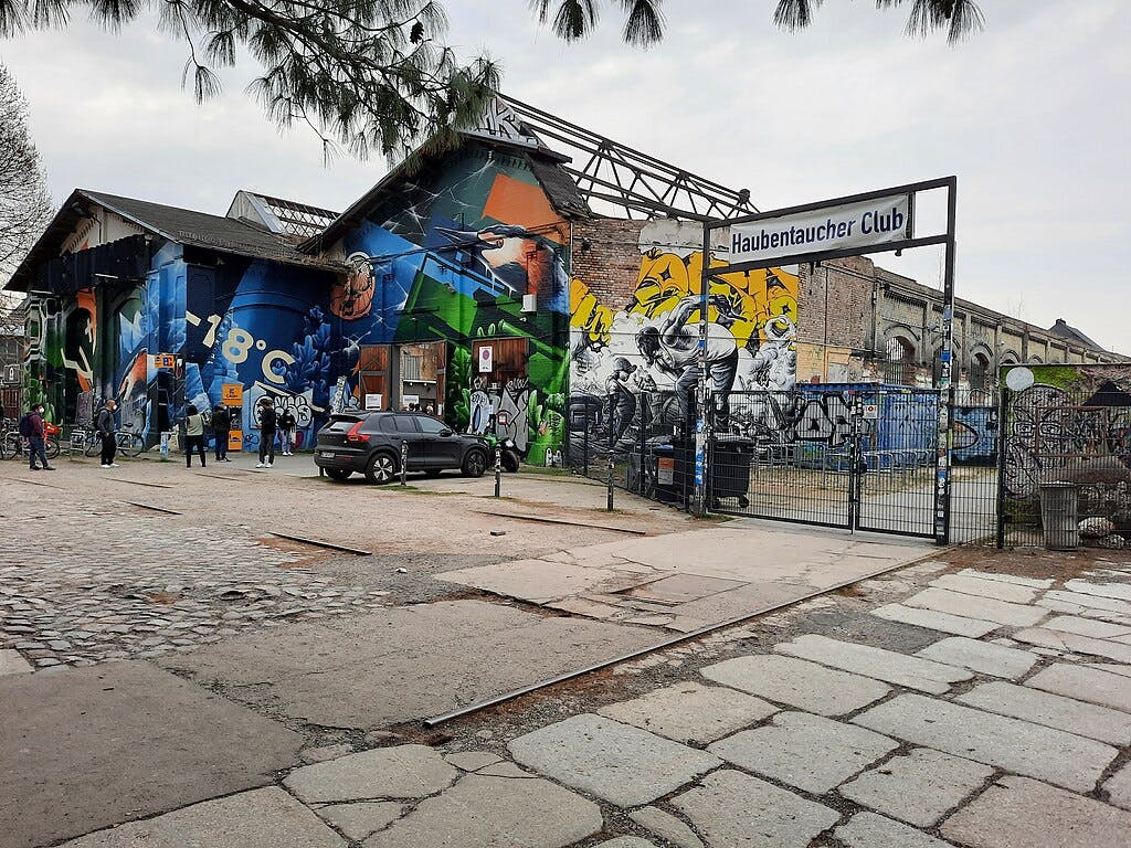 Průmyslová budova pokrytá graffiti s lidmi v okolí a cedulí s nápisem „Haubentaucher Club“ nad bránou.