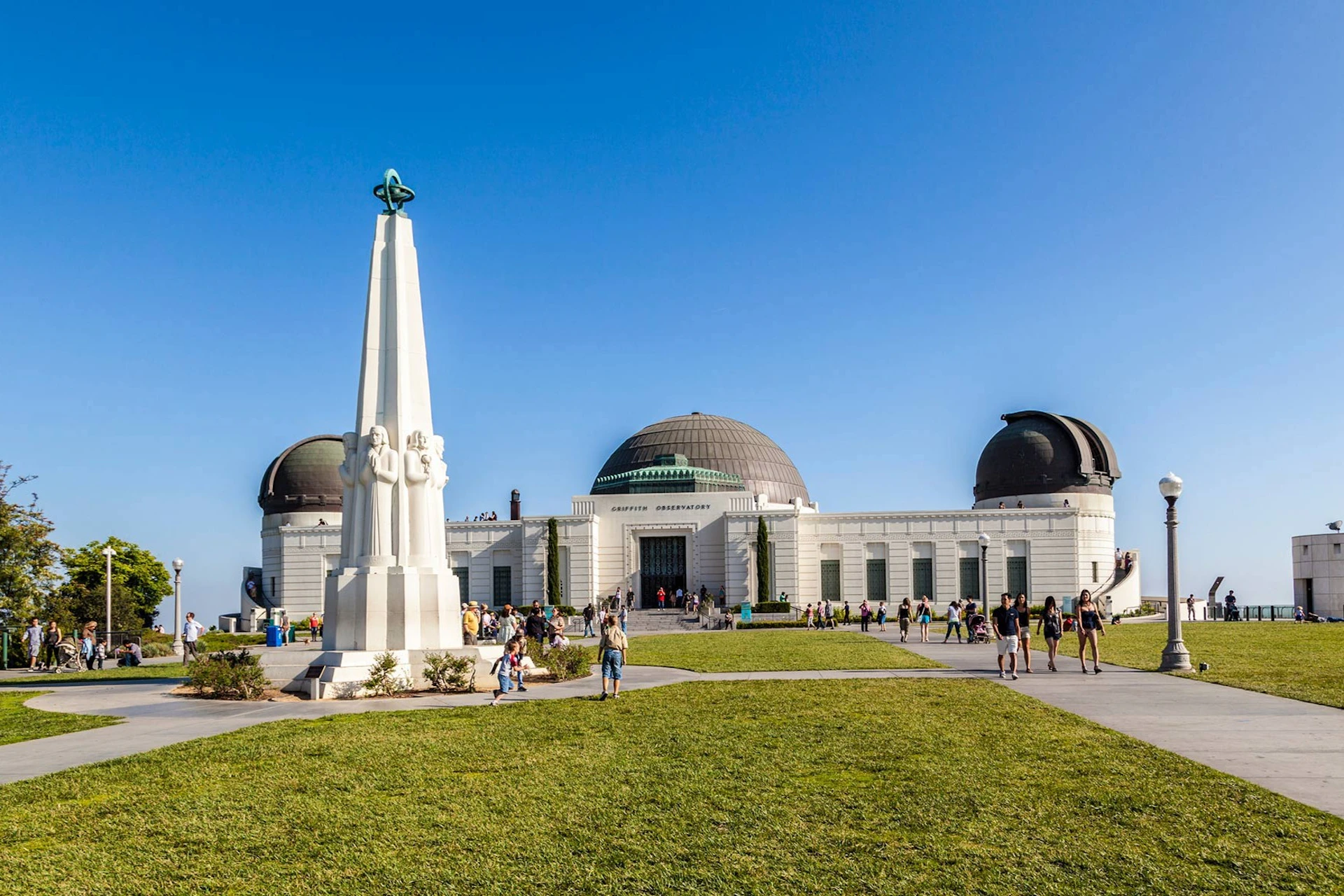 A large white observatory with domes, fronted by a tall obelisk and grassy area filled with people on a clear day.