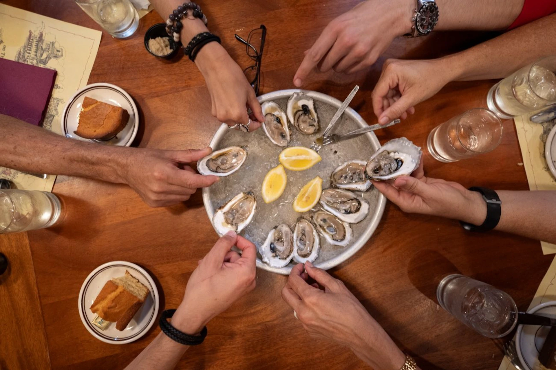 Several hands holding oysters from a plate containing oysters and lemon wedges on a wooden table.