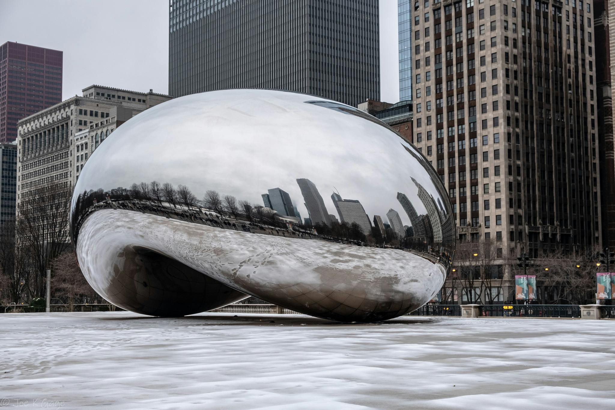 En stor sølvreflekterende skulptur i en bypark med høje bygninger i baggrunden. Jorden er snedækket.