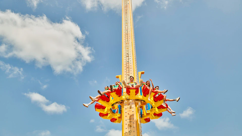 A group of people seated on a colorful amusement park drop tower ride against a blue sky with clouds.