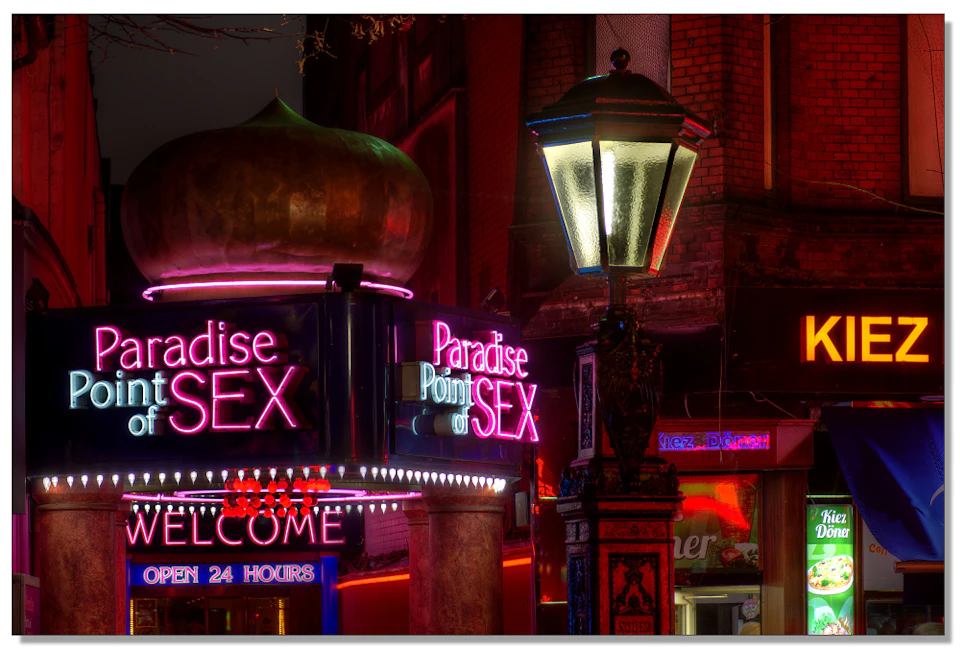 Neon signs for "Paradise Point of SEX" and "WELCOME OPEN 24 HOURS" illuminated in bright pink and purple lights at night.