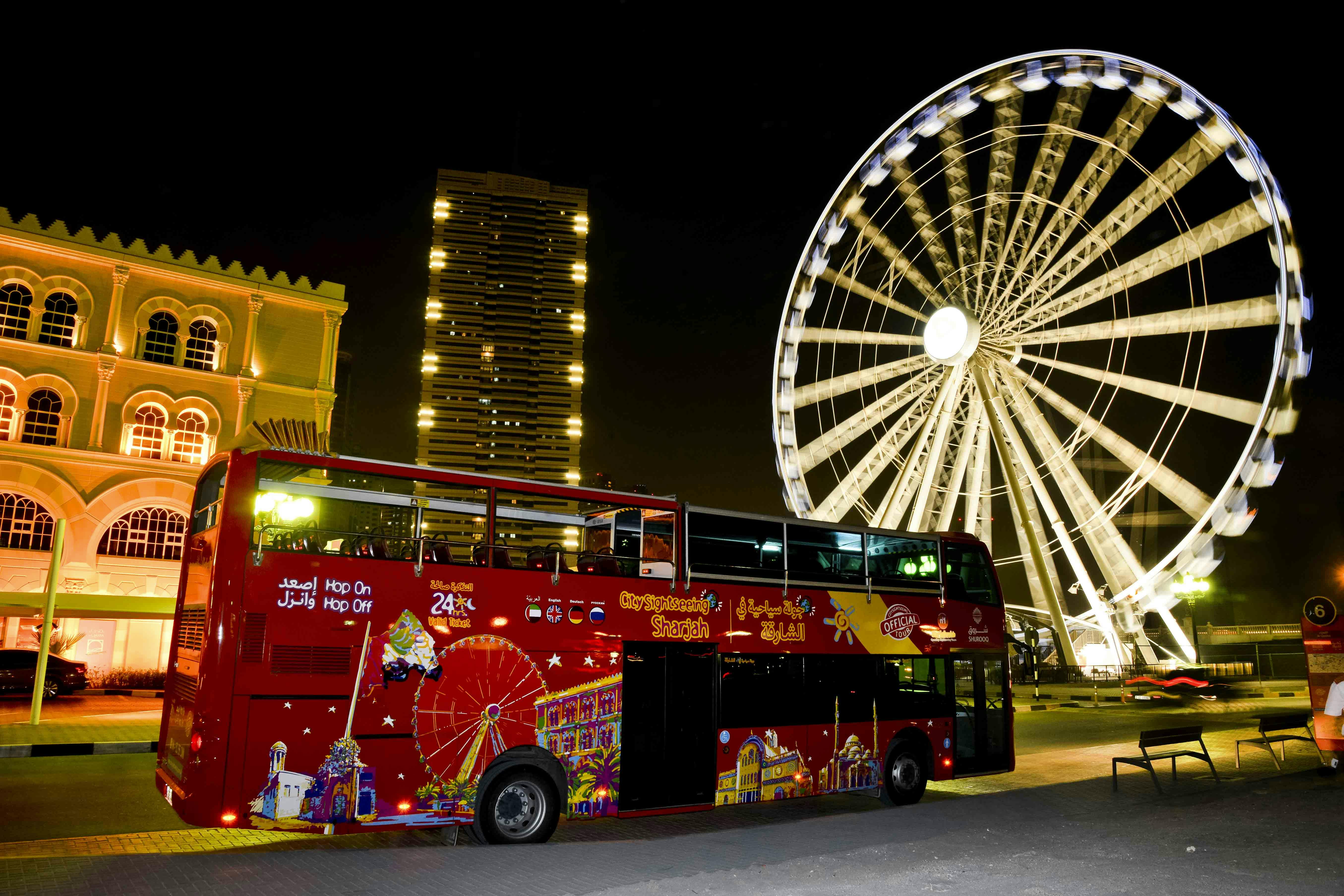 A red double-decker tour bus with city landmarks painted on it is parked near a large, illuminated Ferris wheel at night.