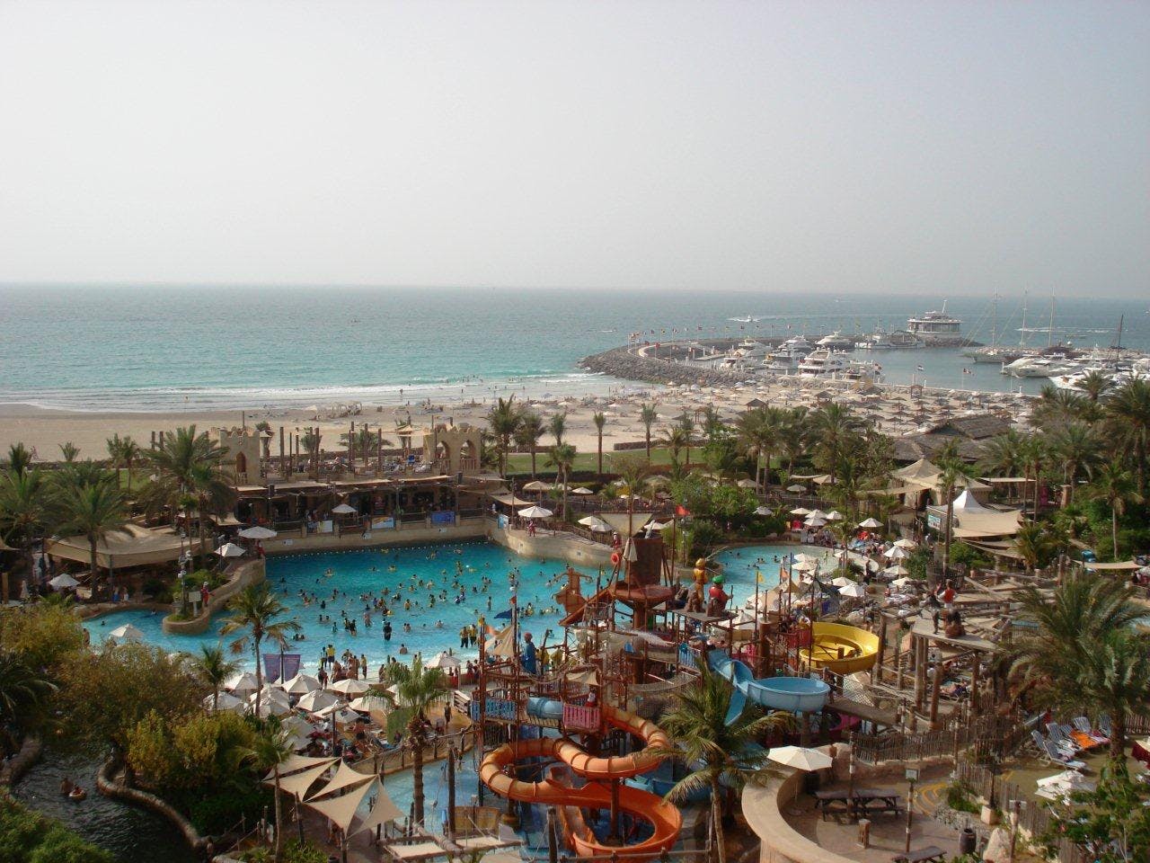 미끄럼틀, 여러 개의 수영장, 야자수, 사람들이 어트랙션을 즐기는 해변가 워터파크의 공중 전망. 바다와 배가 있는 정박지가 배경으로 보입니다.