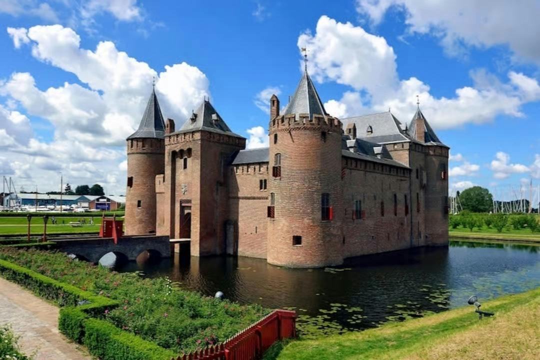 Un castello medievale in mattoni con torri e un fossato, immerso nel verde sotto un cielo azzurro e limpido punteggiato da nuvole soffici.