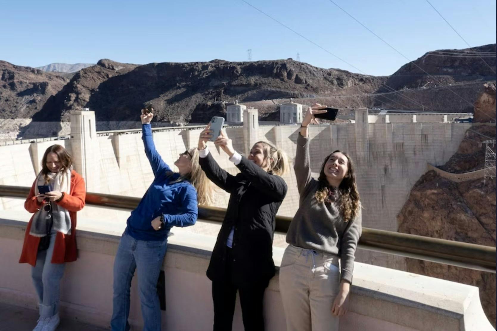 Four people standing on a balcony near a dam, taking photos and enjoying the scenery under a clear blue sky.