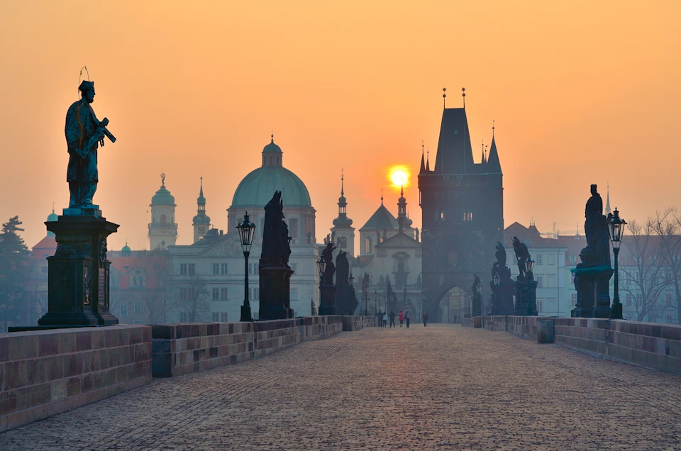 A cobblestone bridge with statues and lampposts, leading to a Gothic tower. People walk towards buildings at sunrise.