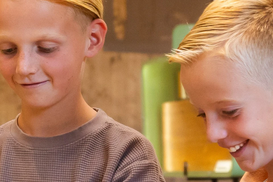 Two boys playing a keyboard in an indoor setting with wooden walls and a display area in the background.