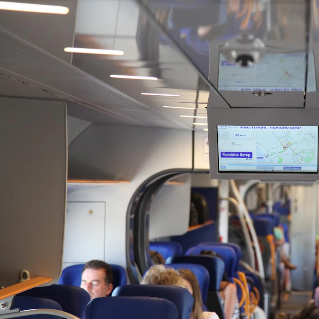 People seated in a modern train with blue seats, overhead screens display information about the train's route and next stops.